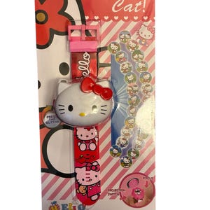 May include: A Hello Kitty watch on a pink and red card. The watch has a white face with a red bow and a red band with Hello Kitty images. The card reads "Hello Cat!" and has a projection switch.