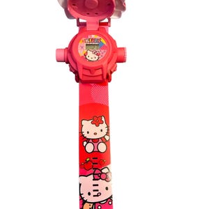 May include: A pink Hello Kitty watch with a white protective cover. The watch face displays the time and a Hello Kitty graphic. The red watch band features Hello Kitty illustrations and apple graphics.