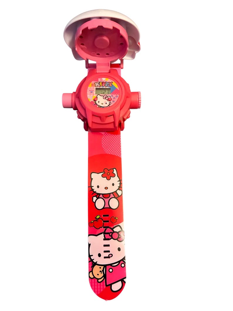May include: A pink Hello Kitty watch with a white protective cover. The watch face displays the time and a Hello Kitty graphic. The red watch band features Hello Kitty illustrations and apple graphics.
