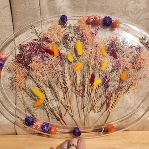Könnte beinhalten: Eine ovale, transparente Harzschale mit Trockenblumen. Die Schale ist mit einer Vielzahl von Trockenblumen in Orange-, Gelb-, Lila- und Burgundertönen gefüllt. Die Blumen sind in einem dekorativen Muster angeordnet.