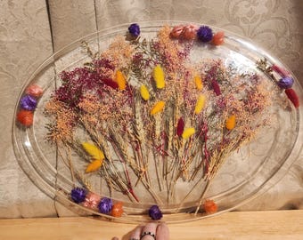 May include: An oval-shaped, clear resin tray with dried flowers. The tray is filled with a variety of dried flowers in shades of orange, yellow, purple, and burgundy. The flowers are arranged in a decorative pattern.