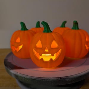 May include: A group of five small, orange, jack-o'-lantern pumpkin decorations with green stems. Each pumpkin features a carved, glowing face with triangular eyes and a jagged mouth, illuminated from within by a warm, soft yellow light.