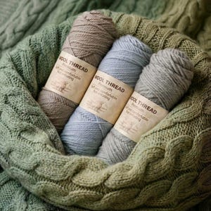 May include: Three skeins of Wool Thread in shades of gray and blue, nestled in a green knitted basket. The yarn labels read "Premium, Soft and comfortable, Not irritating". A green knitted fabric forms the background.