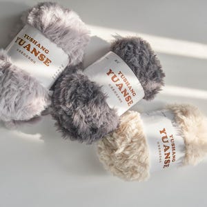 Puede incluir: Tres ovillos de lana esponjosa en tonos grises y crema. Cada ovillo tiene una etiqueta con el texto "YUSHANG YUANSE". La lana parece suave y texturizada, adecuada para proyectos de punto o ganchillo.