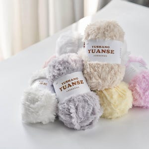Puede incluir: Un montón de ovillos de lana esponjosa en varios colores pastel, incluyendo blanco, gris, beige, amarillo y rosa. Cada ovillo tiene una etiqueta que dice "YUSHANG YUANSE". Ideal para proyectos de tejido y ganchillo.