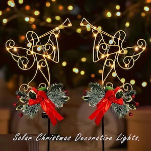 May include: Two solar-powered Christmas decorative lights shaped as angels playing trumpets. The gold angel figures have small, warm white lights. Each angel has a base decorated with faux greenery, red berries, and a red bow. The text "Solar Christmas Decorative Lights" is at the bottom.