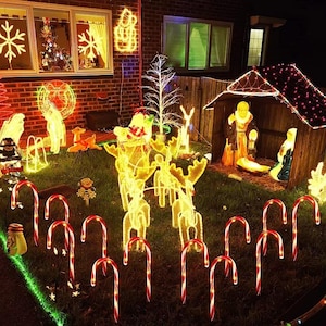 Puede incluir: Una exhibición navideña nocturna al aire libre con renos iluminados, bastones de caramelo y un pesebre. El jardín está decorado con varias luces festivas, incluyendo copos de nieve y un árbol de Navidad en la ventana.