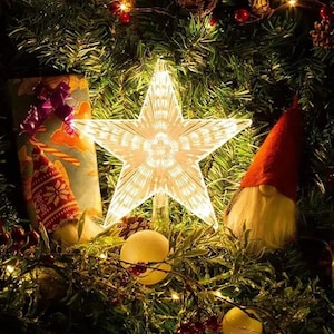 Puede incluir: Una estrella iluminada para la parte superior del árbol de Navidad. La estrella es blanca y tiene una superficie texturizada. La estrella está rodeada de agujas de pino verdes, bayas rojas y adornos decorativos. Una escena festiva.