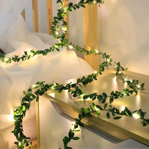 May include: A decorative string of lights with small, bright bulbs intertwined with green artificial leaves. The garland is draped across a wooden surface and a white sheet, creating a warm, inviting ambiance. The leaves are a deep green color.