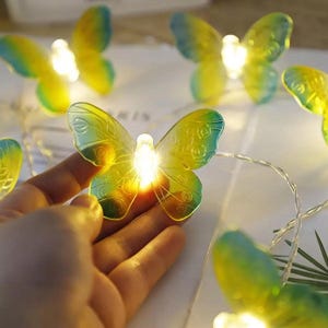 Pode incluir: Uma série de luzes decorativas em forma de borboleta. Cada borboleta é amarela e verde com contorno azul e contém uma pequena luz. As luzes são presas em um fio fino e prateado, criando um ambiente caprichoso.