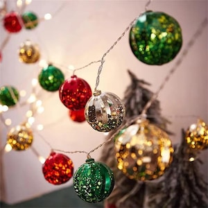 Può includere: Una stringa di luci natalizie con decorazioni a sfera in vetro rosso, verde, oro e argento. Gli ornamenti sono rotondi e hanno una finitura glitterata e riflettente. Le luci sono infilate su un sottile filo d'argento.