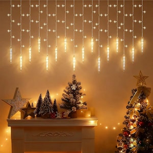 Puede incluir: Una escena navideña cálida y acogedora con una repisa de chimenea adornada con árboles de Navidad en miniatura, adornos de estrella y velas. Encima, luces de carámbano y luces en forma de estrella iluminan la habitación. Un árbol de Navidad decorado está a la derecha.