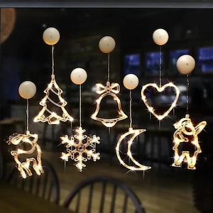 May include: A collection of illuminated Christmas-themed decorations, including a Christmas tree, bell, heart, reindeer, snowflake, moon, and a unicorn. Each decoration is outlined with warm white lights and suspended from a small, round, white light.