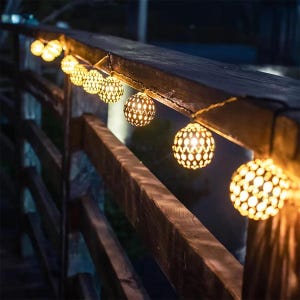 Op de afbeelding: Decoratieve lichtslinger met bolvormige, patroonvormige bollen die een warm licht uitstralen. De lichten zijn langs een houten reling geregen en creëren een gezellige sfeer. Perfect om een vleugje charme toe te voegen aan tuinen of binnenruimtes.