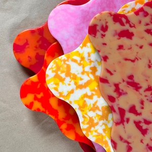 Puede incluir: Una pila de cinco posavasos de bordes ondulados con varios patrones jaspeados. Los colores incluyen naranja brillante, rosa sólido, un diseño moteado en blanco y amarillo, y una pieza beige con manchas de color rojo oscuro, dispuestos en abanico.