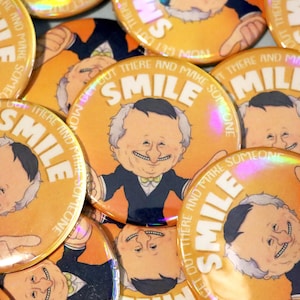 Puede incluir: Un conjunto de botones naranjas con una ilustración de dibujos animados de un hombre sonriente con traje y pajarita. La palabra "SMILE" se muestra de forma destacada, con la frase "GET OUT THERE AND MAKE SOMEONE" alrededor.