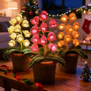 Peut inclure: Trois arrangements floraux décoratifs et illuminés dans des pots tressés. Les fleurs sont crochetées en jaune, rouge et orange, avec des lumières internes. Des feuilles et des tiges vertes complètent le design. Les arrangements sont posés sur une table en bois, avec un sapin de Noël et des décorations en arrière-plan.