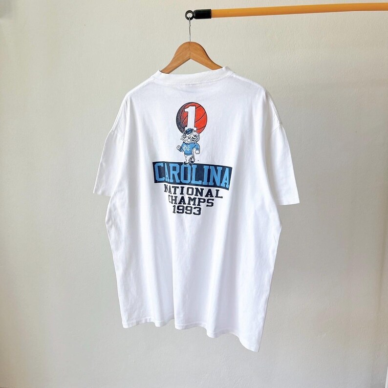 May include: White t-shirt with a graphic on the back featuring the word "CAROLINA" in blue, the text "NATIONAL CHAMPS 1993", and a cartoon character holding a basketball. The shirt is hanging on a wooden hanger.