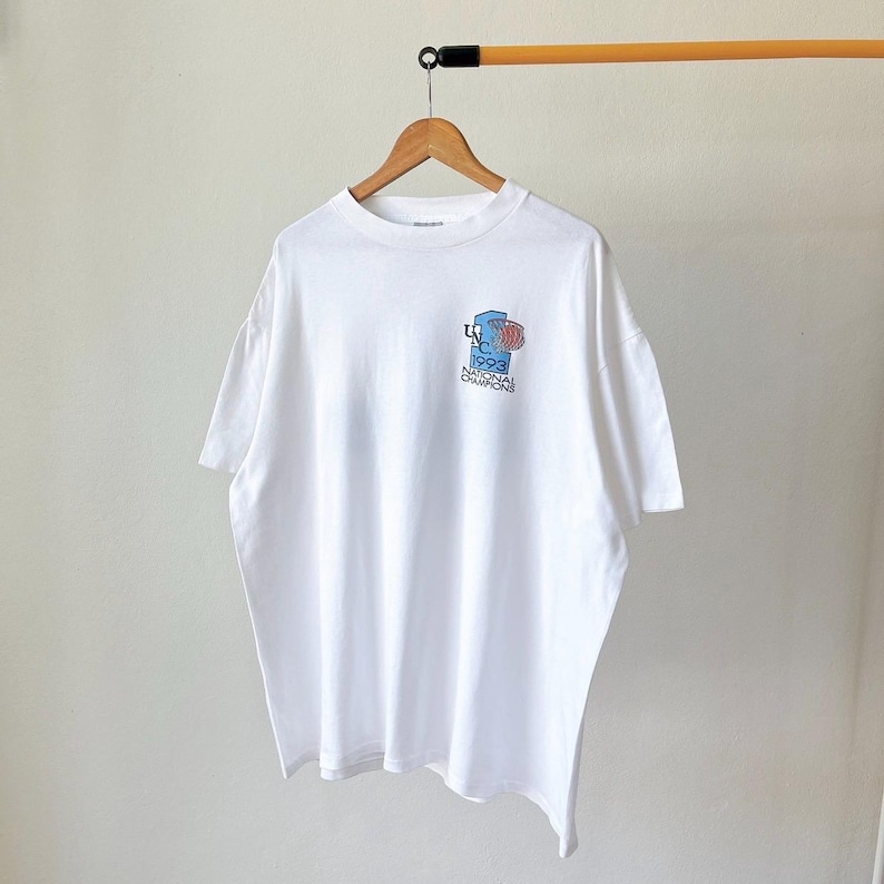 May include: White t-shirt with a small graphic on the front. The graphic features a basketball hoop and the text "UNC 1993 National Champions" in blue, red, and white. The shirt is hanging on a wooden hanger.