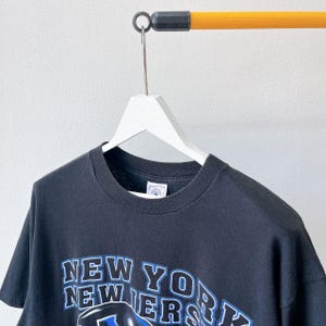 Puede incluir: Camiseta negra con el texto "NEW YORK NEWERS" en letras blancas y azules. La camiseta est&aacute; colgada en una percha blanca contra una pared blanca. La camiseta est&aacute; hecha de un material suave.
