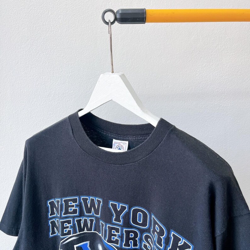 Puede incluir: Camiseta negra con el texto "NEW YORK NEWERS" en letras blancas y azules. La camiseta est&aacute; colgada en una percha blanca contra una pared blanca. La camiseta est&aacute; hecha de un material suave.