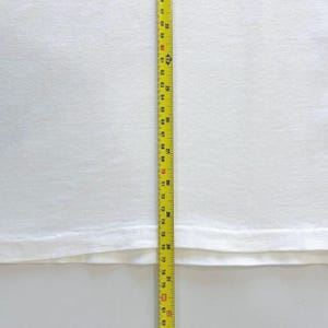 May include: A white fabric, possibly a shirt or sheet, is measured with a yellow and black tape measure. The tape measure shows measurements from 57 to 82 inches. The fabric is plain and smooth.