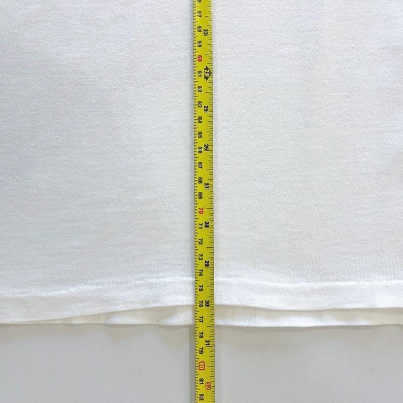 May include: A white fabric, possibly a shirt or sheet, is measured with a yellow and black tape measure. The tape measure shows measurements from 57 to 82 inches. The fabric is plain and smooth.
