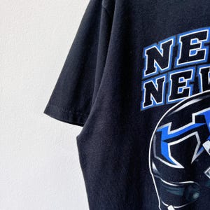 Puede incluir: Camiseta negra con el texto "NE" en blanco con contorno azul. La camiseta presenta un gr&aacute;fico de un casco con detalles en azul y blanco. La camiseta est&aacute; hecha de una tela oscura y suave.