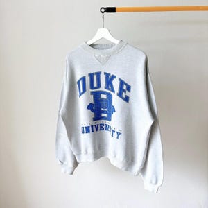 May include: A light gray crewneck sweatshirt featuring the blue text DUKE UNIVERSITY and a large letter D logo on the chest. The garment has long sleeves, a ribbed collar with a V-shaped insert, and ribbed cuffs and hem, hanging on a white hanger.