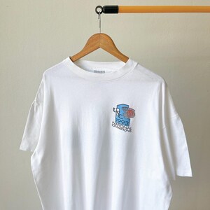 May include: White t-shirt with a small graphic on the front. The graphic features a basketball hoop and the text "UNC 1993 National Champions" in blue and red. The shirt is hanging on a wooden hanger.