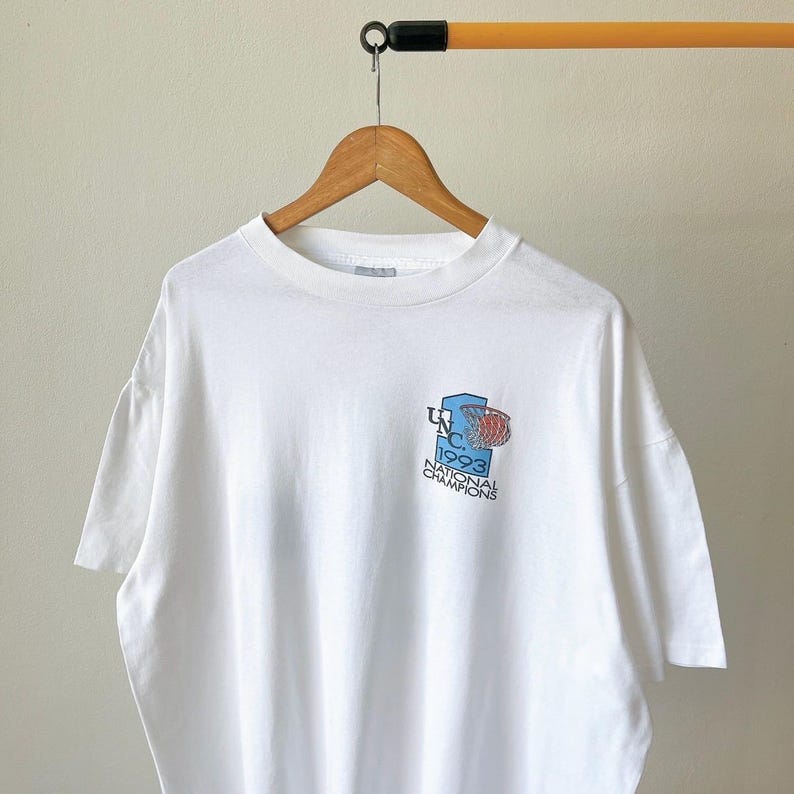 May include: White t-shirt with a small graphic on the front. The graphic features a basketball hoop and the text "UNC 1993 National Champions" in blue and red. The shirt is hanging on a wooden hanger.