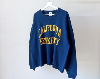 May include: A navy blue crewneck sweatshirt featuring the text CALIFORNIA BERKELEY in yellow block lettering across the chest. The garment has long sleeves, a ribbed collar, and a small white brand tag visible at the inner neckline.