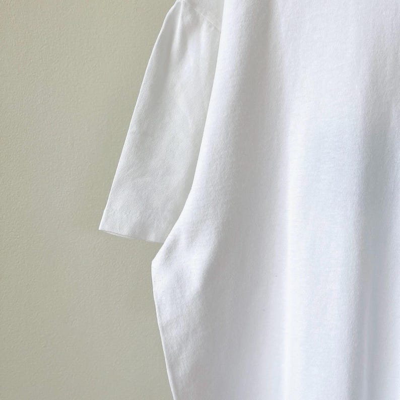 May include: A close-up shot of a white t-shirt, showcasing the fabric's texture and the design of the short sleeve. The shirt is plain and appears to be made of a soft, breathable material. The background is a neutral, light-colored wall.