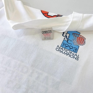 May include: White XXL t-shirt featuring a graphic commemorating the UNC 1993 National Champions. The design includes a basketball going into a net, with the text "UNC 1993 NATIONAL CHAMPIONS". The shirt has a crew neck and a tag that reads "ONEITA POWER-T".