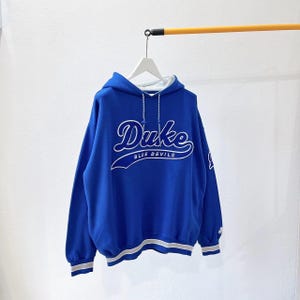 Może przedstawiać: Królewska niebieska bluza z kapturem z napisem "Duke" w białym skrypcie na piersi, z napisem "BLUE DEVILS" poniżej. Bluza ma biały sznurek, białą podszewkę w kapturze oraz biało-szare paski na mankietach i na dole.
