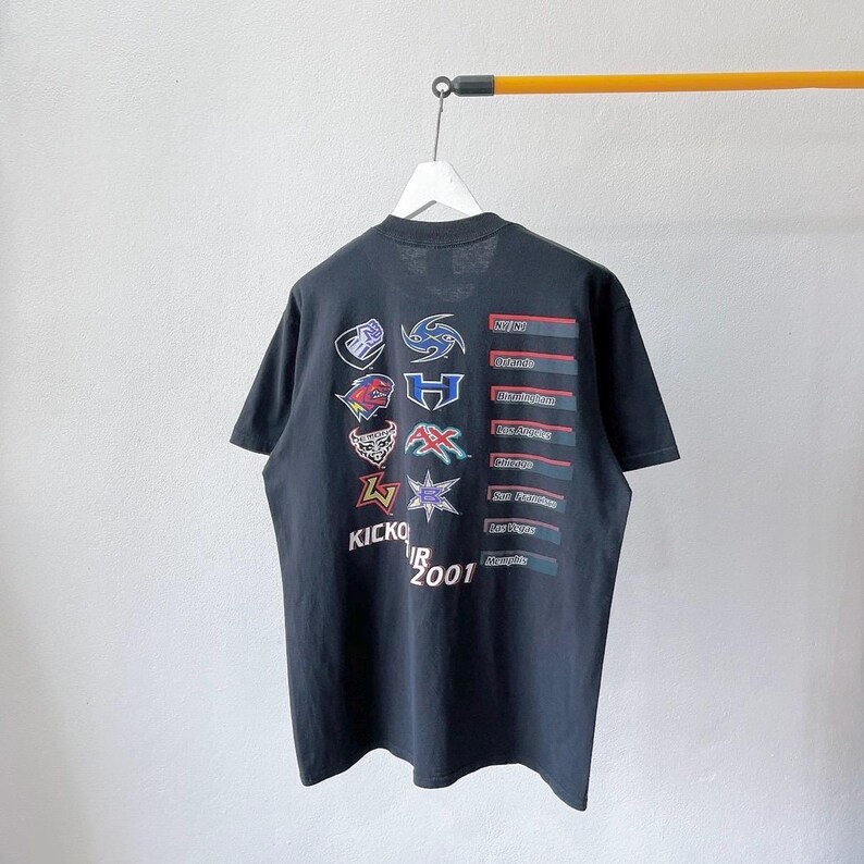 Puede incluir: Camiseta negra con m&uacute;ltiples logotipos y texto. La camiseta muestra varios logotipos de equipos y las palabras "KICKOFF TOUR 2001". La camiseta est&aacute; colgada en una percha blanca sobre un fondo liso.