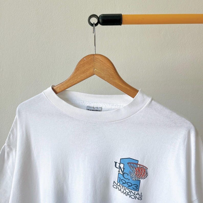 May include: White crew neck t-shirt with a graphic featuring a basketball net and the text "UNC 1993 National Champions" in blue and red. The shirt is hanging on a wooden hanger.