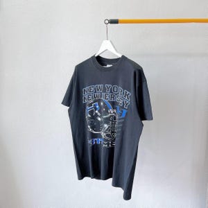 Puede incluir: Camiseta negra con un dise&ntilde;o de casco de f&uacute;tbol y las palabras "NEW YORK NEW JERSEY" en blanco y azul. La camiseta tiene cuello redondo y mangas cortas. El dise&ntilde;o incluye el texto "5TH AVE" y "MIE".
