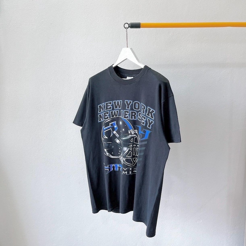 Puede incluir: Camiseta negra con un dise&ntilde;o de casco de f&uacute;tbol y las palabras "NEW YORK NEW JERSEY" en blanco y azul. La camiseta tiene cuello redondo y mangas cortas. El dise&ntilde;o incluye el texto "5TH AVE" y "MIE".