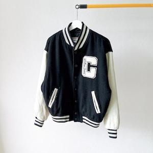May include: A black varsity jacket with cream-colored sleeves, featuring a large white letter C patch on the left chest with the text College Field written inside. The jacket has a button-front closure, striped ribbed cuffs, and a striped ribbed collar.