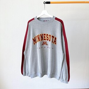 Op de afbeelding: Een grijze sweatshirt met het woord "MINNESOTA" in kastanjebruine en gouden letters. De sweatshirt heeft kastanjebruine strepen langs de mouwen en het woord "GOPHERS" staat eronder. Het hangt aan een witte hanger.