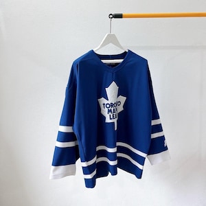 May include: A blue hockey jersey with white stripes on the sleeves and hem. The jersey displays a white maple leaf logo with "TORONTO MAPLE LEAFS" printed on it. The jersey is suspended from a white hanger.