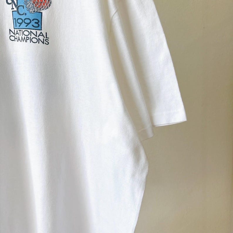 May include: White t-shirt with a graphic featuring a basketball, the text "N.C. 1993" and "NATIONAL CHAMPIONS" in black and blue. The shirt is made of a soft, white fabric.