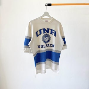 May include: White short-sleeve shirt with blue and gray stripes. The shirt features the text "UNA" in blue arched lettering above a circular emblem and the word "WOLPACK" below. The shirt is hanging on a white hanger.