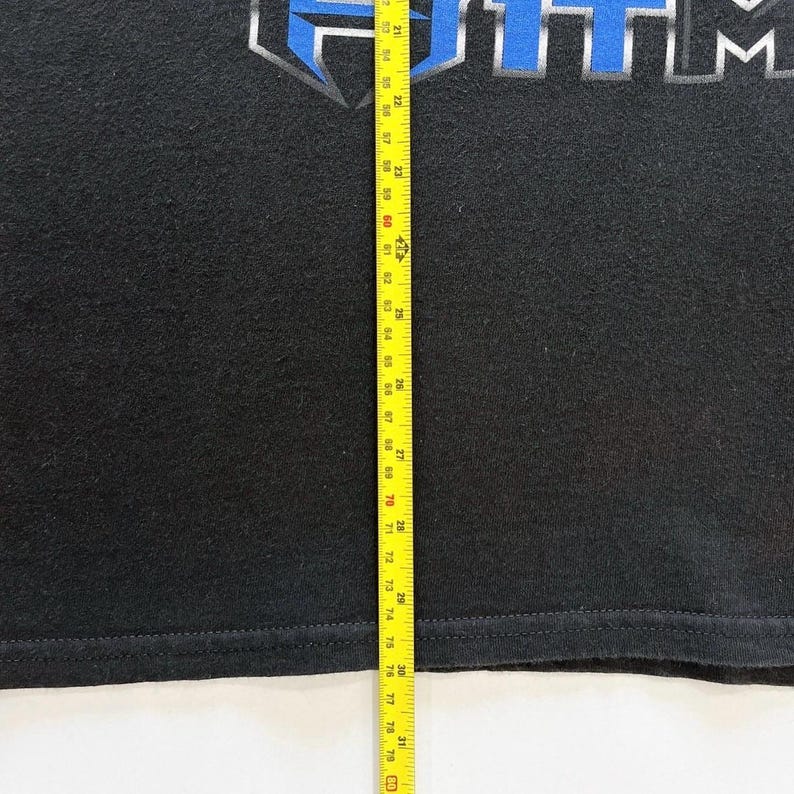 Puede incluir: Camiseta negra con la palabra "ITM" en un dise&ntilde;o azul y plateado. Una cinta m&eacute;trica amarilla se coloca verticalmente a lo largo de la camiseta, mostrando medidas de 53 cm a 200 cm.