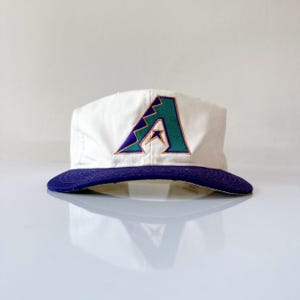 Pode incluir: Boné de beisebol branco com aba roxa e logotipo "A" bordado em verde-azulado e roxo. O boné tem uma coroa estruturada e uma aba curva.