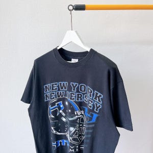 Puede incluir: Camiseta negra con un dise&ntilde;o de casco de f&uacute;tbol y el texto "NEW YORK NEW JERSEY" en blanco y azul. La camiseta est&aacute; colgada de una percha blanca sobre un fondo neutro.