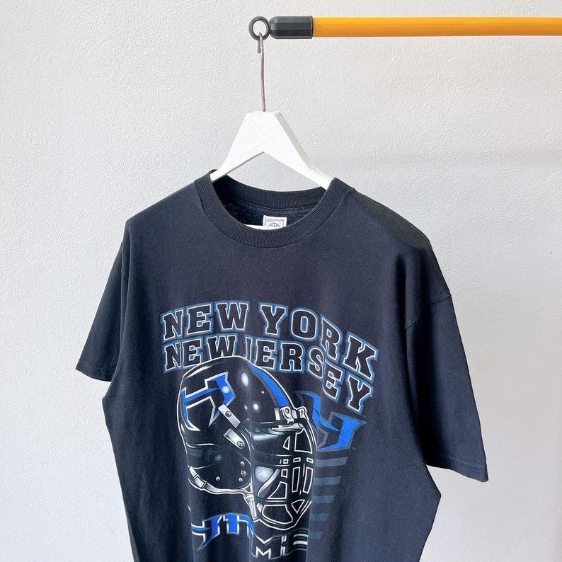 Puede incluir: Camiseta negra con un dise&ntilde;o de casco de f&uacute;tbol y el texto "NEW YORK NEW JERSEY" en blanco y azul. La camiseta est&aacute; colgada de una percha blanca sobre un fondo neutro.