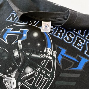 Puede incluir: Camiseta negra con un gr&aacute;fico de casco de f&uacute;tbol en azul y blanco, y el texto "NEW JERSEY". La camiseta tiene un aspecto vintage, con una etiqueta Delta Pro Weight. El dise&ntilde;o es una representaci&oacute;n gr&aacute;fica llamativa de un tema deportivo.
