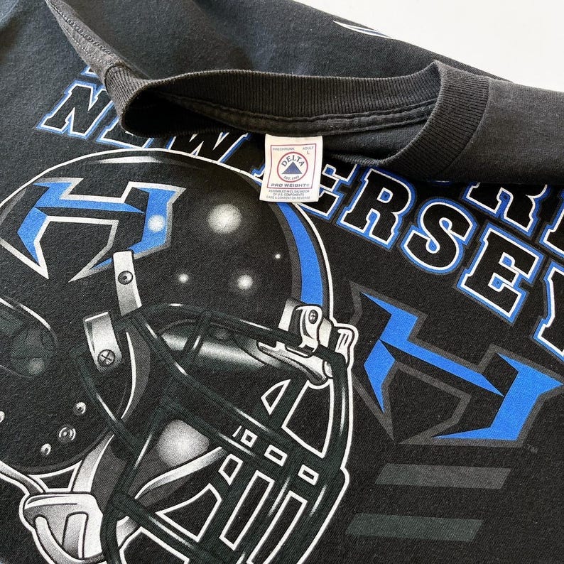 Puede incluir: Camiseta negra con un gr&aacute;fico de casco de f&uacute;tbol en azul y blanco, y el texto "NEW JERSEY". La camiseta tiene un aspecto vintage, con una etiqueta Delta Pro Weight. El dise&ntilde;o es una representaci&oacute;n gr&aacute;fica llamativa de un tema deportivo.